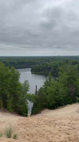 Scenic view from a sandy dune overlooking a forested lake on a cloudy day. A calm, overcast landscape of water, trees, and sky.