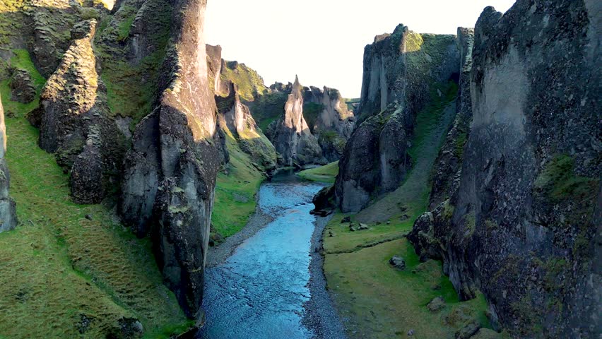 Experience the breathtaking beauty of Fjadrargljufur Canyon in Iceland, where towering cliffs rise majestically above a tranquil river, surrounded by vibrant green landscapes.