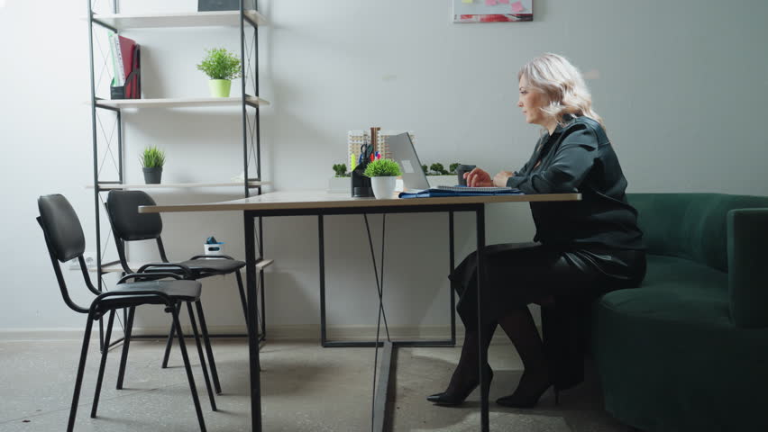 Sales woman seated in stylish office multitasks by answering customer phone call while working on laptop, surrounded by organized desk items, real estate model