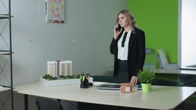 real estate agent seated in modern office on phone call with client opening laptop to start work surrounded by architectural model, stationery, green plant, and organized files on shelf in background - Powered by Shutterstock - Get 15% off with code: PIKWIZARD15