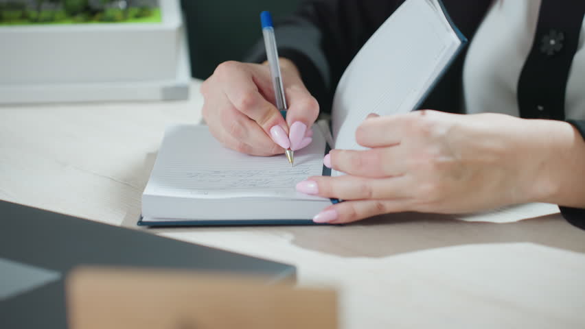 close up of adult woman with long pink nails taking down handwritten notes in lined journal while seated at desk with open laptop and architectural building model slightly visible in background