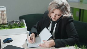 businesswoman multitasking at desk holding phone between ear and shoulder while writing in notebook with polished nails, working in bright modern office with architectural model in background - Powered by Shutterstock - Get 15% off with code: PIKWIZARD15