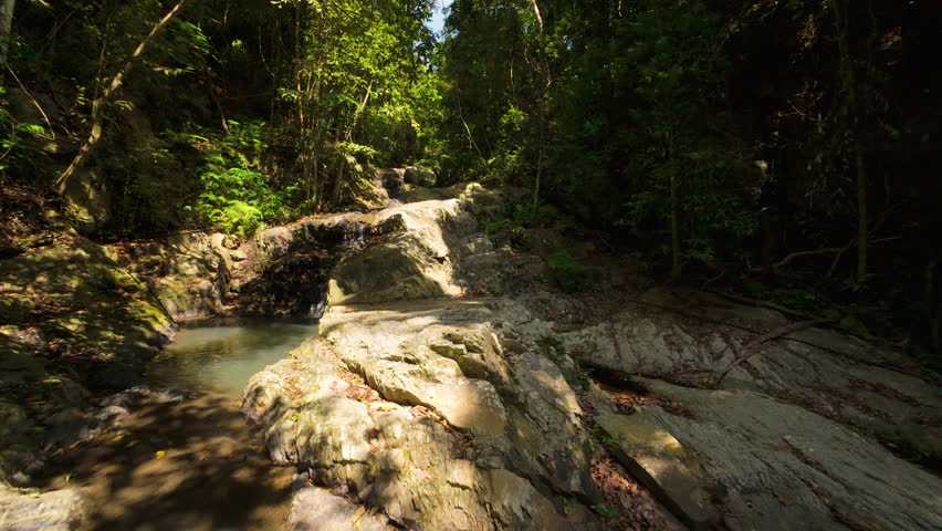 FPV Drone Flying Low Above Shallow Rocky Stream with Small Waterfall and Dense Jungle Surrounding in Bright Daylight.