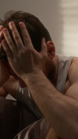 Vertical shot of depressed bearded man sitting on bed and hitting head hysterically suffering from panic attacks