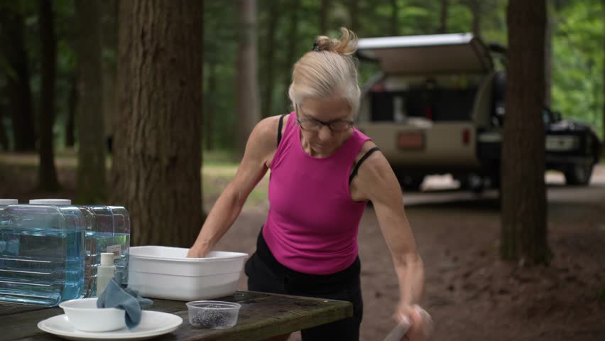 A mature retired woman is washing dishes outside at a campground surrounded by trees, with a teardrop camper trailer parked nearby. Enjoying a simple outdoor lifestyle.