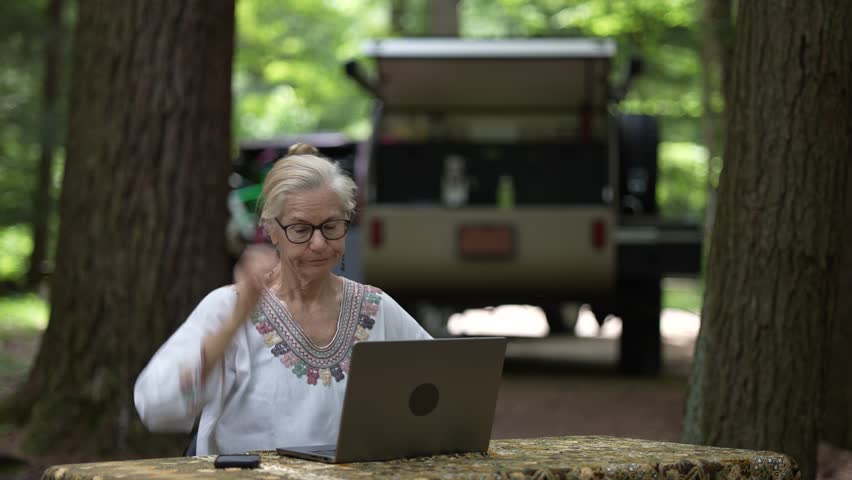 Frustrated, unhappy mature woman sits at a table in a forest campground, using a computer and smartphone while a teardrop camper trailer is visible in the background.