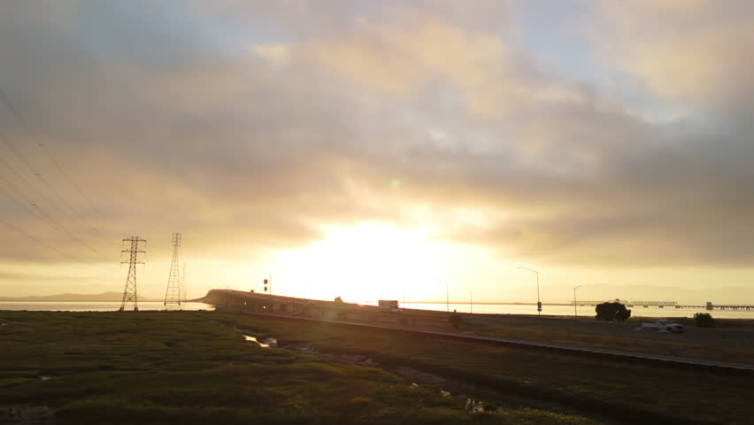 Powerful pan left shot showing transmission tower and Dumbarton Bridge with soft sunrise light over wetlands in summer.