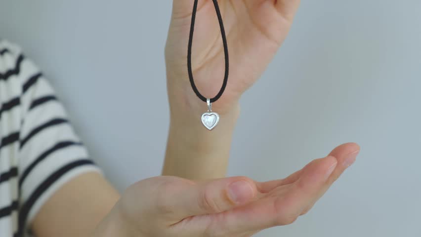 A person is holding a necklace with a heart pendant. The necklace is black and silver. The person is wearing a white shirt
