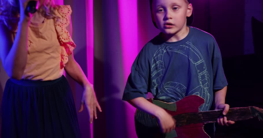 energetic kids stage concert performance at home. boy playing cardboard guitar and girl sing in the microphone. neon lighting. music band