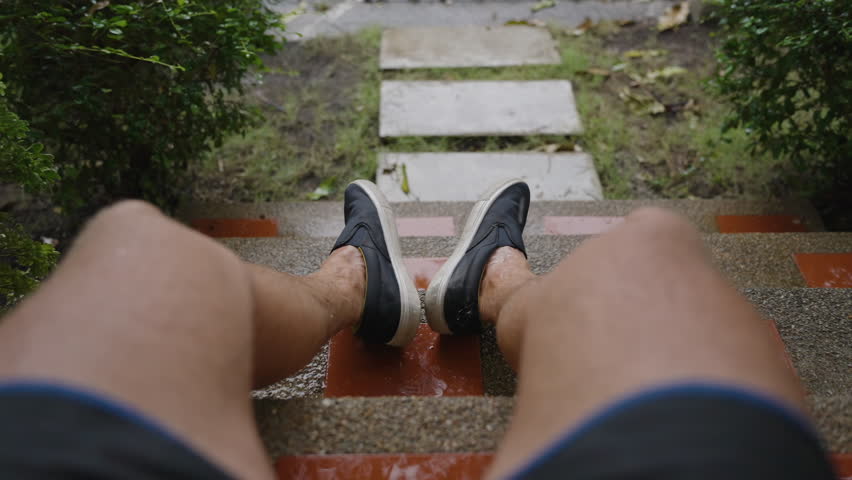 Close-Up First-Person View of Man Sitting on Rainy Steps with Legs Wet from Tropical Downpour and Garden Path Ahead in Slow Motion, Rainy Mood Concept.