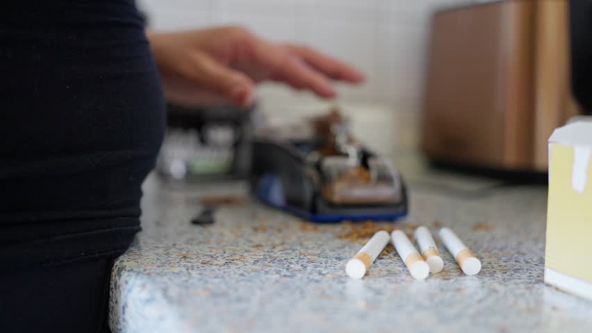 A person is using a machine to roll cigarettes while having tobacco scattered across the kitchen counter. Slow motion