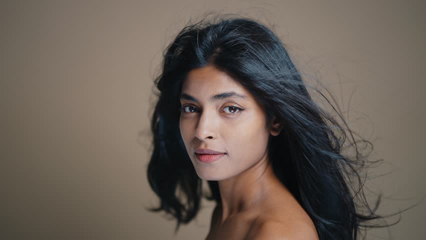 Professional studio portrait capturing young indian woman with long dark hair expressing genuine emotions while posing against neutral brown background. 