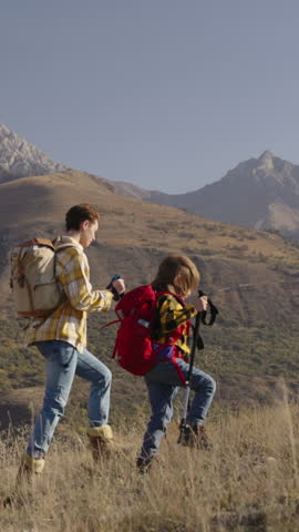 Mother and child hike autumn mountain path with trekking poles. Colorful leaves and golden sunlight create peaceful scene of active family adventure. Slow motion shows joy of exploring nature together