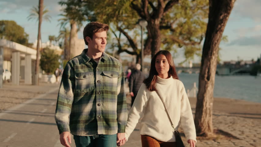 A young couple holds hands and walks along the Guadalquivir River in Seville, Spain, on a sunny day. They are happy and in love.