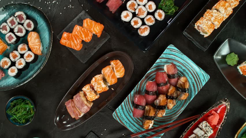 Top view of traditional japanese cuisine . Sushi rolls set with salmon , tuna fish and vegetable served on black stone table . Asian food on black stone slate with chopsticks . Camera movement .