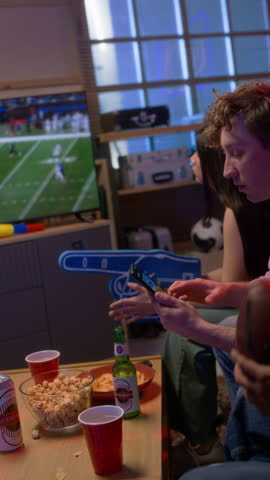 Vertical shot of multiethnic group of friends watching sports match on TV together and cheering joyfully jumping up, man holding smartphone with betting app on screen