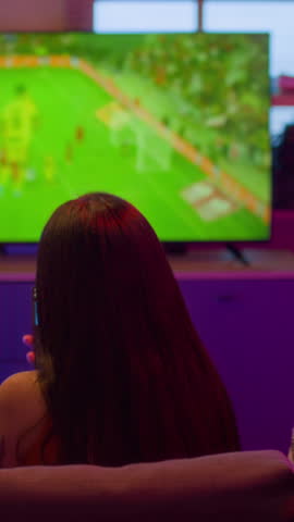 Vertical back view shot of three sports fans sitting on couch together watching football match on TV and cheering