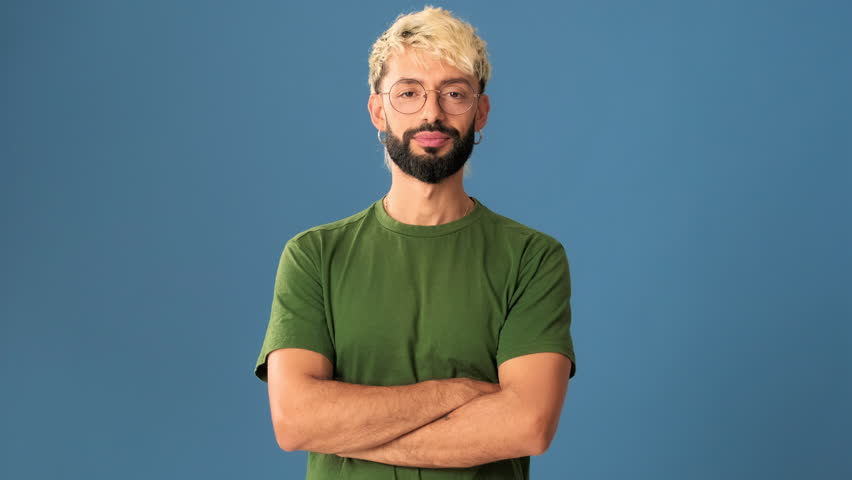 A guy with glasses wearing an olive t-shirt crossing his arms and looking at the camera with a smile isolated on blue background in a studio