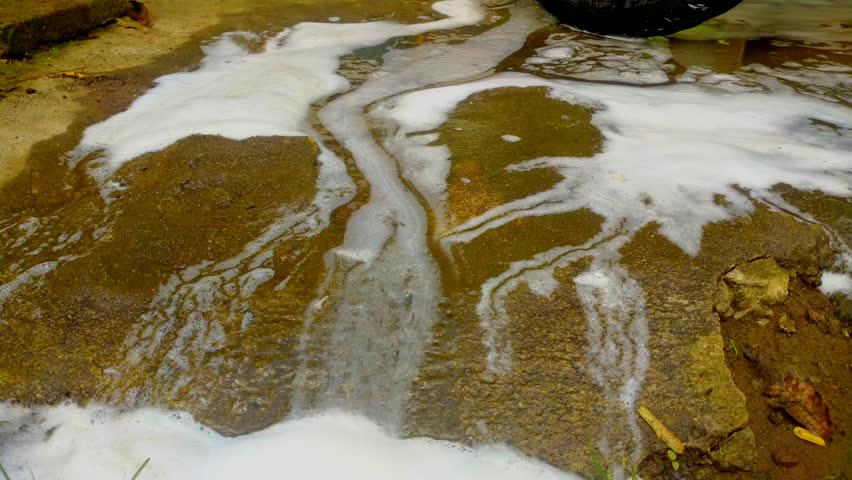 White, thick soap suds and murky water flow across a textured, dark concrete surface, with a small patch of green grass visible at the bottom.