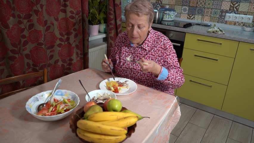 An elderly woman eats fish and vegetables on a plate with a knife and fork. A grandmother is having lunch at home in the kitchen. Healthy eating. Bananas on table