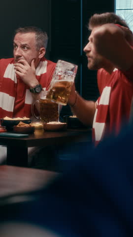 Vertical shot of young male friends in red uniform having beer while sitting by table in pub and watching football broadcast with tense facial expression
