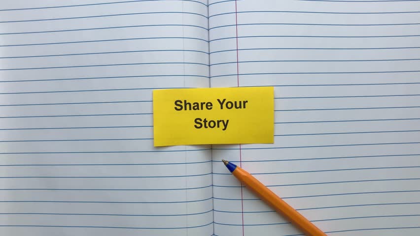 A yellow paper note with the words Share Your Story on it lying in an open notebook near a ballpoint pen. Close up.