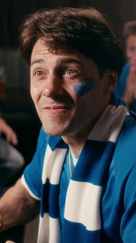 Vertical shot of football fans in blue t-shirts watching broadcast in pub, then standing up, embracing each other, shouting and jumping excitedly after their favorite team scoring goal