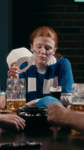 Vertical shot of young unhappy female football fan touching her head while sitting by table with snacks and glasses of beer after watching match broadcast