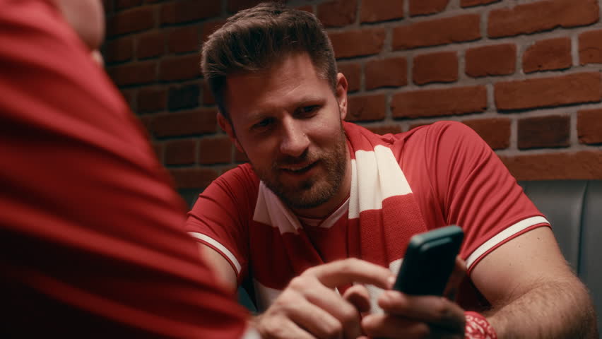 Two young male and female football fans wearing red sportswear sitting in front of camera in pub and discussing online game in smartphone while pointing at screen