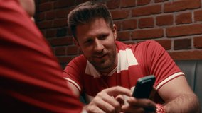 Two young male and female football fans wearing red sportswear sitting in front of camera in pub and discussing online game in smartphone while pointing at screen - Powered by Shutterstock - Get 15% off with code: PIKWIZARD15