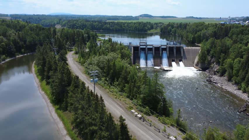 A view of a hydroelectric dam with cascading water and full-flow spillway gates, combining industrial design with a lush forest setting, showcases sustainable renewable energy production. Mitis-2 Dam.