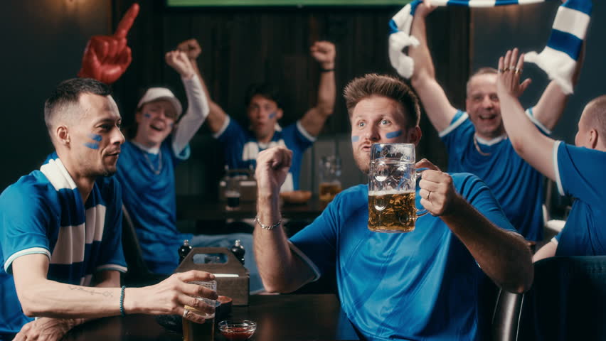 Two guys in blue t-shirts sitting by table in pub and discussing latest news while watching football match broadcast against group of tense friends expressing joy after goal of their favorite team
