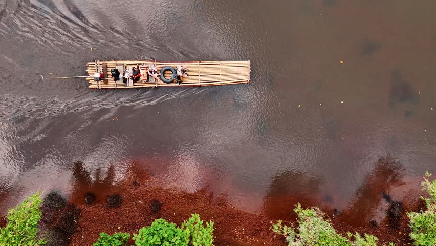 Travelers rafting bamboo raft across serene river, drifting through verdant tropical scenery during sunny day in traditional southeast asian waterway exploration, Thailand