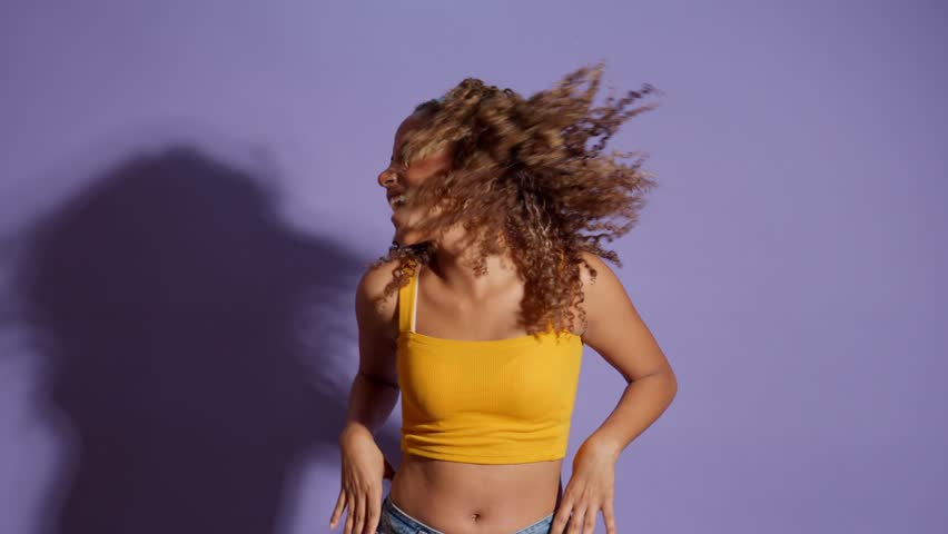Happy young african american woman with curly hair enjoys dancing and flipping her hair in slow motion, isolated on a vibrant purple background