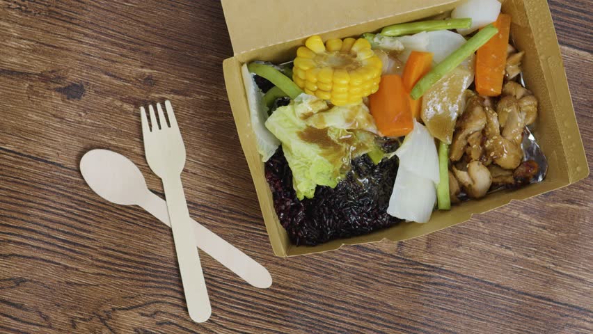Disposable wooden cutlery is placed near a cardboard takeaway box containing black rice, teriyaki chicken, corn on the cob, carrots, green beans, bok choy and daikon radish