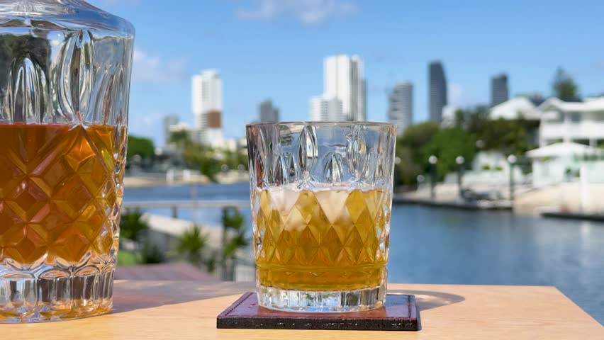 A hand lifts a crystal whiskey glass with ice from a wooden table, set against a bright, sunny waterfront city skyline. Static camera, natural daylight