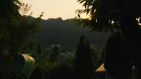 A beautiful trucking shot reveals a golden hour sunset with a sunburst shining through tree leaves, overlooking a peaceful mountain resort nestled in a valley. - Powered by Shutterstock - Get 15% off with code: PIKWIZARD15