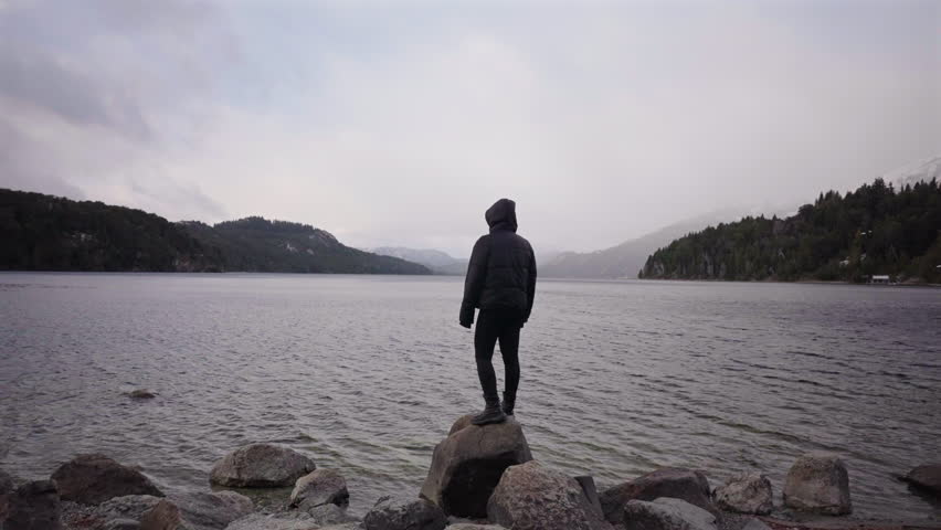 Woman stands alone on rocks facing the misty waters of lake, stillness, healing, introspection