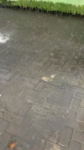 A close-up shot capturing a torrential downpour as large raindrops intensely hit a wet, grey paved surface. The water accumulates into puddles, creating visible ripples and splashes with each drop.
