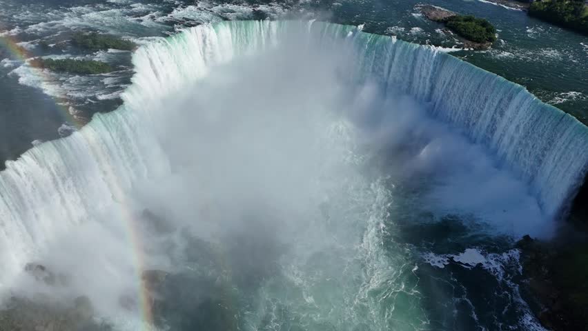 Fixed aerial shot capturing the full curve of Horseshoe Falls from above, with a vibrant rainbow, blue sky, and powerful water currents cascading from the edge to the base