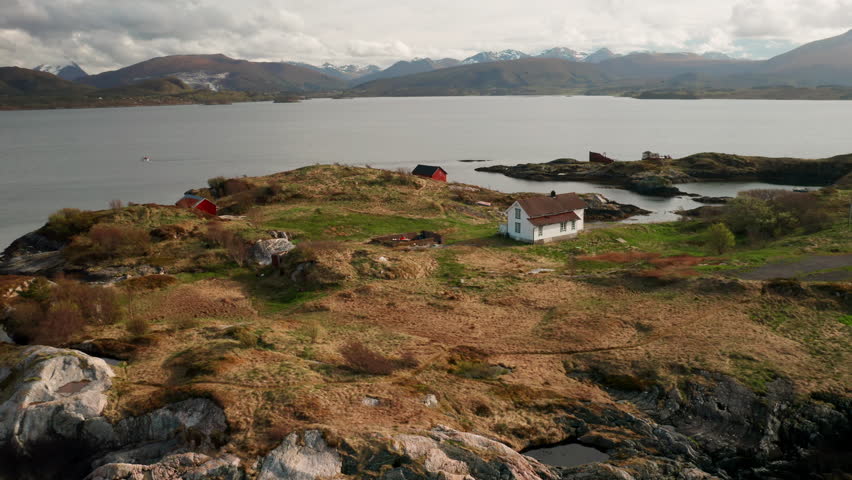 Small house nestled on rocky brown islet among expansive fjord waters of Norwegian coast. Mountains highlight dramatic natural beauty of Norway