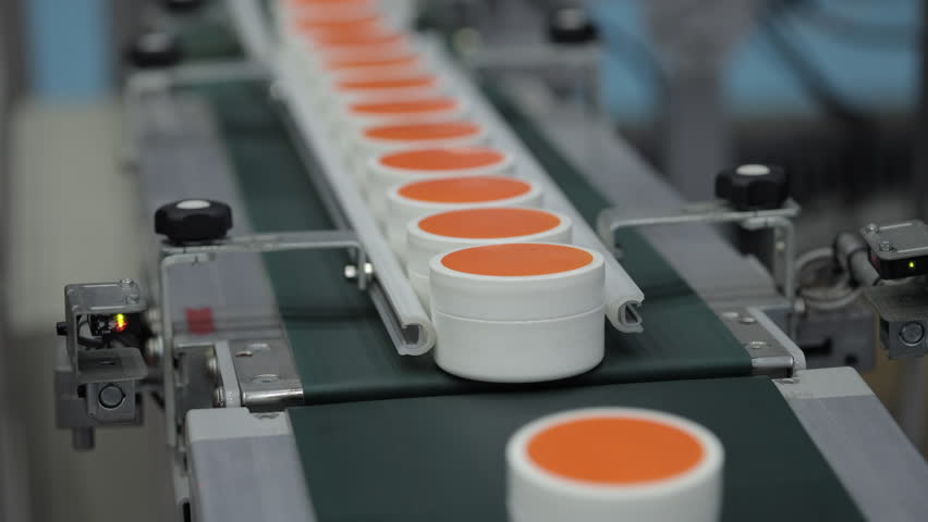 Orange-capped plastic jars with cosmetic cream traveling in a row on an automated conveyor belt in a factory setting, illustrating packaging process in industrial production.