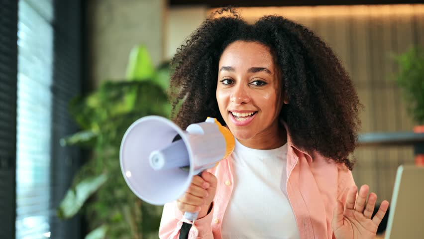 Excited African American woman holding a megaphone and speaking enthusiastically in a modern office setting. Girl appears happy and confident, making an important announcement or sharing news.