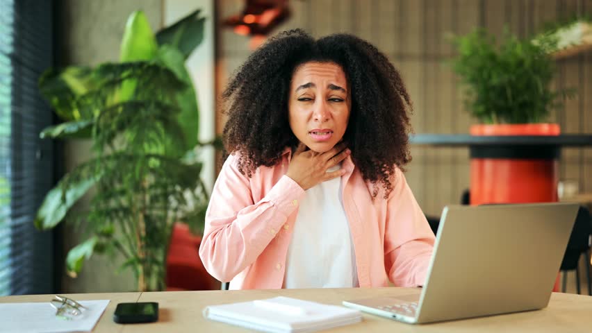 Upset African American woman wearing a pink shirt experiences a painful sore throat while working on her laptop at an office desk. She feels unwell and struggles with discomfort, needing rest.