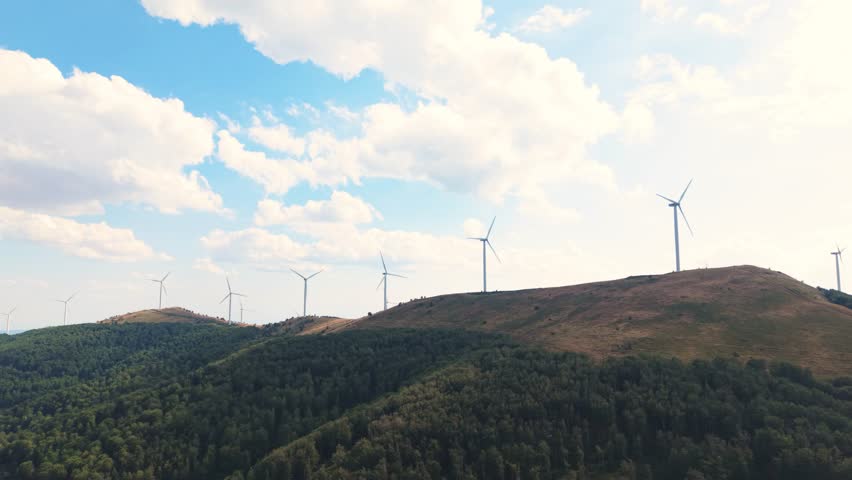 Drone view capturing the movement of renewable energy turbines under a beautiful sky. Mountains and nature offer a peaceful, sustainable backdrop.