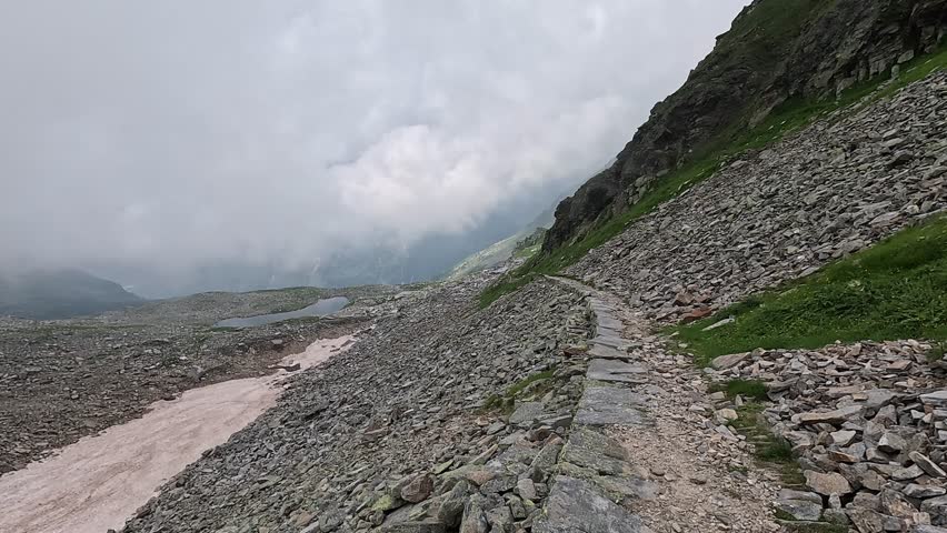 Stone hiking path winds through rugged alpine terrain at Colle del Turlo under a cloudy mountain sky.