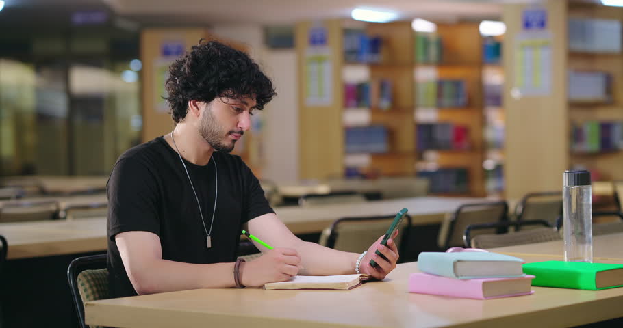 Indian young adult gen z student sitting on desk table use mobile phone hold pencil take online notes write on notebook. Serious smart teenager boy do self study prepare new exam topics indoor library