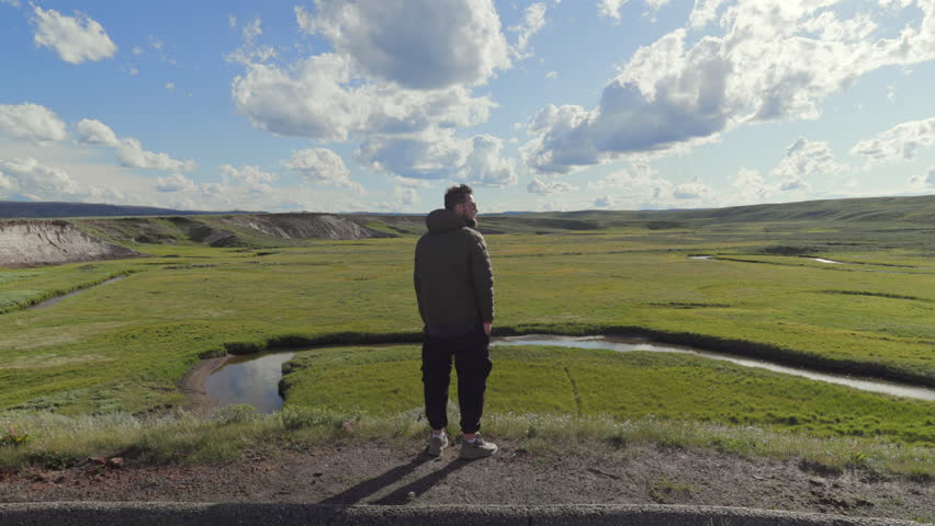 Male tourist stands at a scenic overlook at lamar valley at Yellowstone, gazing at the lush green fields and serene waters below, under a clear blue sky with fluffy clouds, beauty of nature