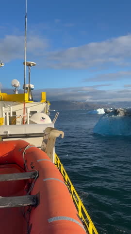 A boat with a life raft drifting near icebergs in the waters of Greenland, with a view of the cloudy sky and mountains in the distance, tc01