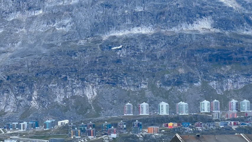 Colorful buildings at the foot of mountains in Greenland with an airplane flying above the rocky landscape.  
